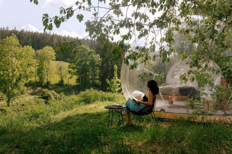 A guest reading in a chair beside the bubble tent, looking out over green hills and forest